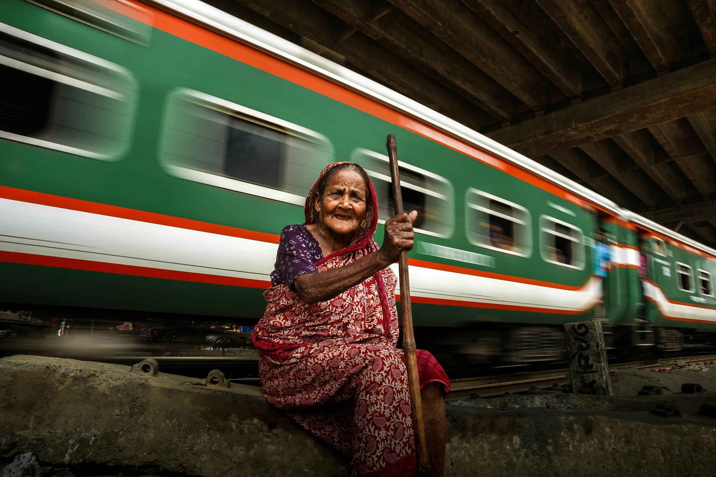 An elderly woman in a red patterned sari sits on a concrete ledge holding a wooden stick, with a green and white train moving past in the background.